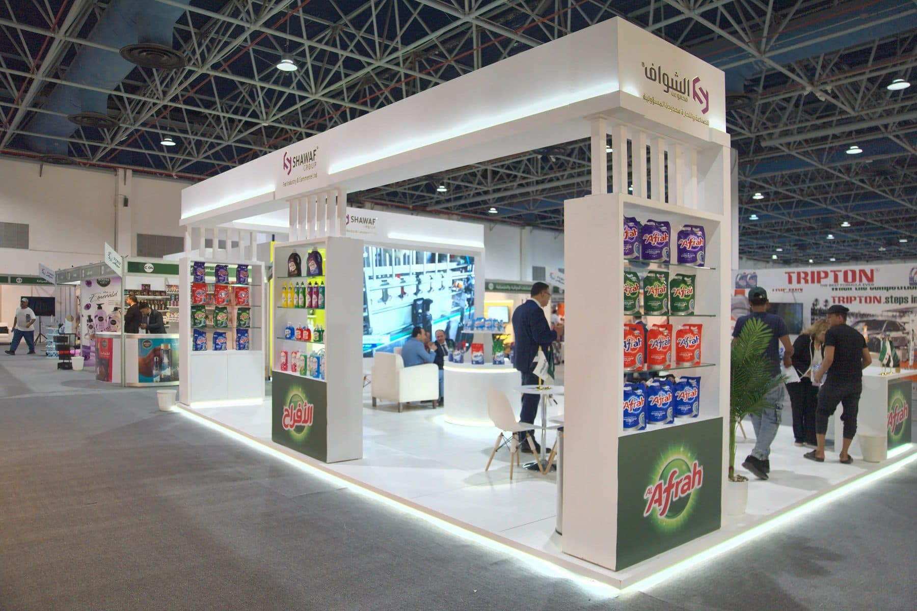Open-corner island exhibition booth built for Shawaf Group with illuminated brand fascia and tiered product shelving